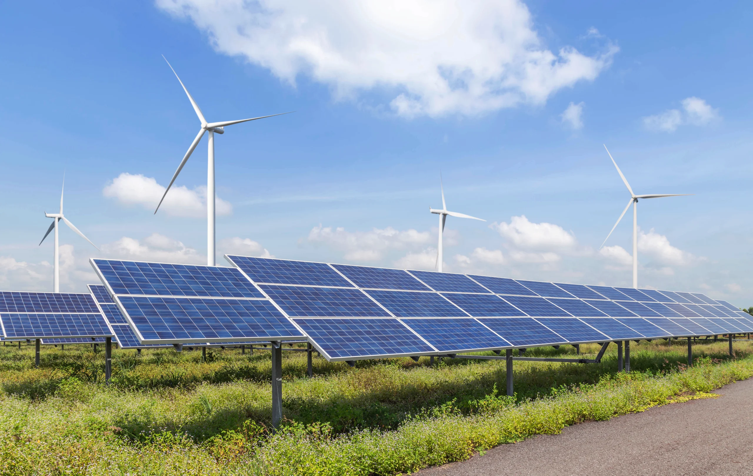 Foto eines Solarparks mit mehreren Reihen blauer Photovoltaikmodule auf einer Grünfläche. Im Hintergrund sind vier Windräder vor blauem Himmel mit weißen Wolken zu sehen.