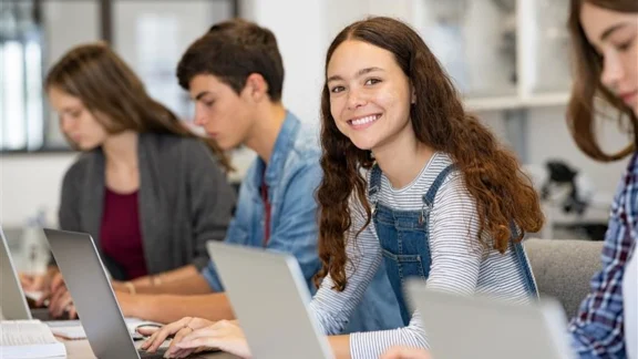 Eine Schülerin lächelt während sie an ihrem Laptop arbeitet. Um sie herum sitzen weitere Schüler*innen, die ebenfalls an deren Laptop arbeiten.