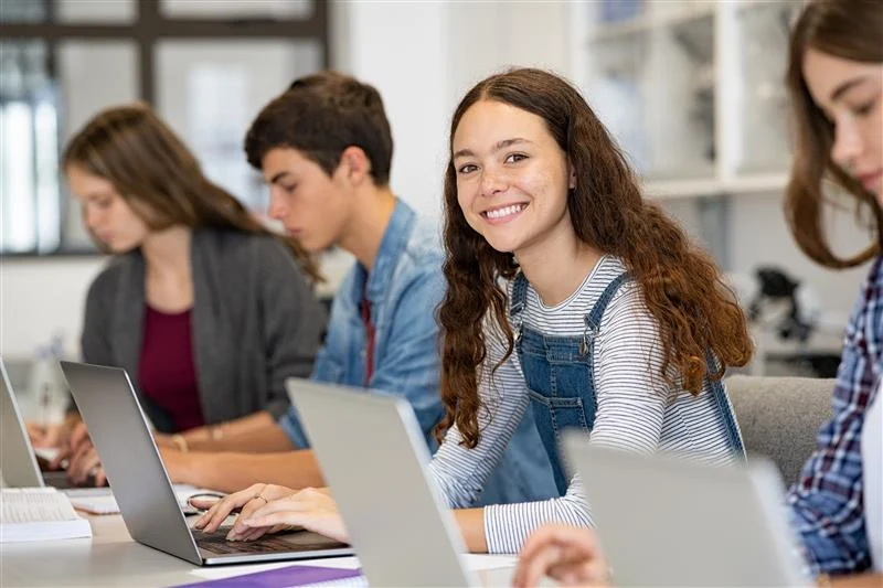 Eine Schülerin lächelt während sie an ihrem Laptop arbeitet. Um sie herum sitzen weitere Schüler*innen, die ebenfalls an deren Laptop arbeiten.