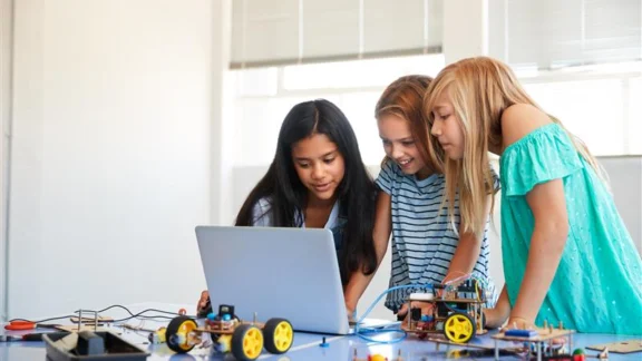 Drei Mädchen im Alter zwischen 8 und 11 Jahren schauen auf den Bildschirm eines Laptops. Vor ihnen liegen Prototypen eines kleinen Elektrofahrzeugs.