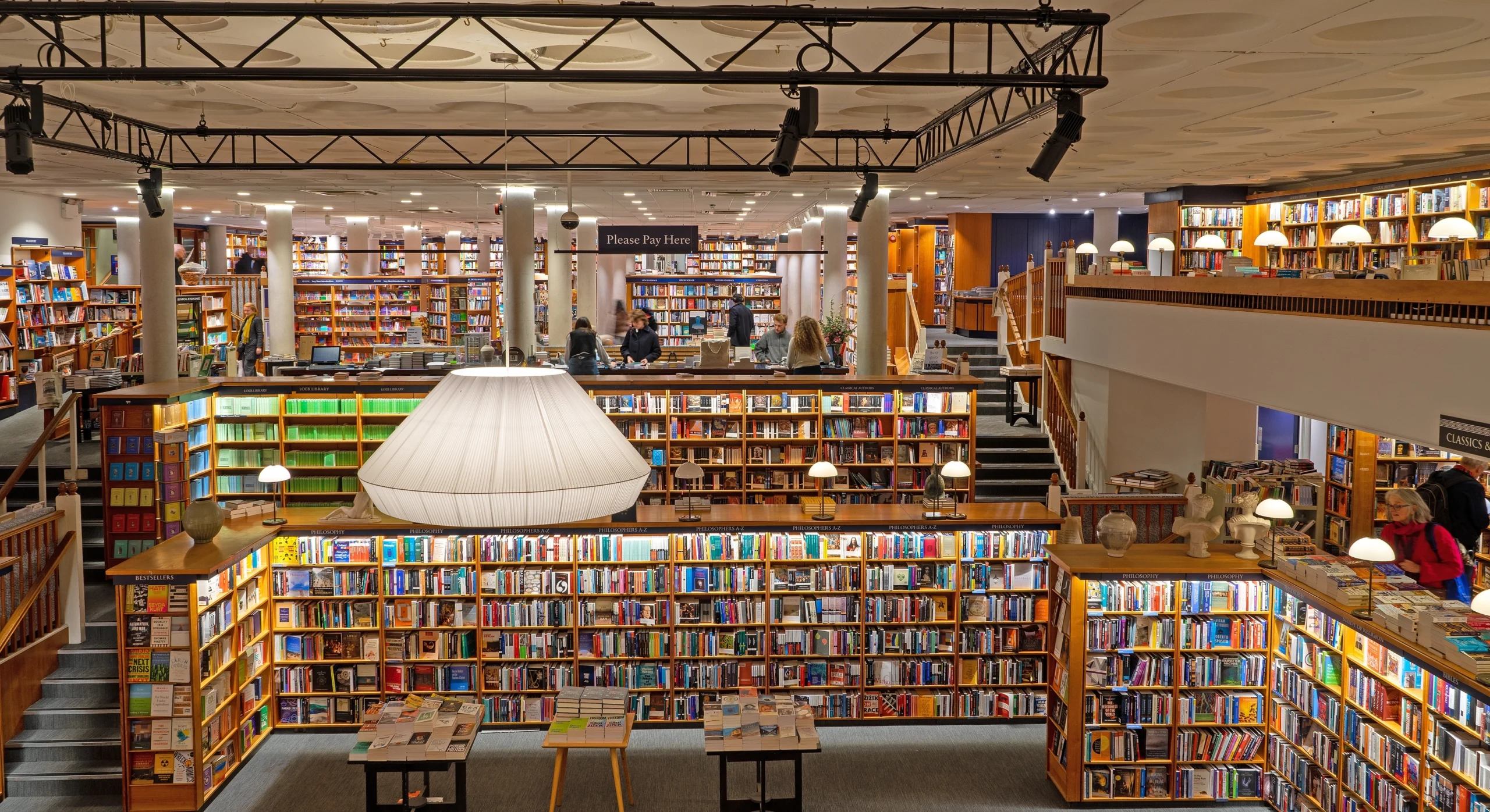 Auf dem Bild ist ein großzügiger Bücherladen mit zwei Etagen zu sehen. Die Regale haben eine Holzoptik und in der Mitte des Raumes hängt eine etwa 2 Meter breite Lampe.