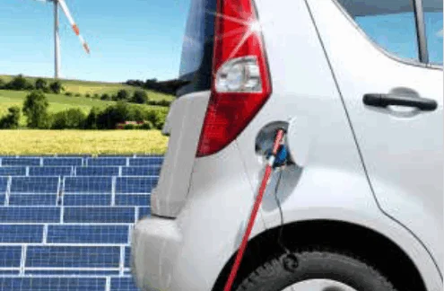 Ein kleines, silbernes Auto wird mit Strom durch ein Kabel versorgt. Im Hintergrund sieht man Solarpanel auf einem offenen Feld sowie ein Windrad bei Sonnenschein.