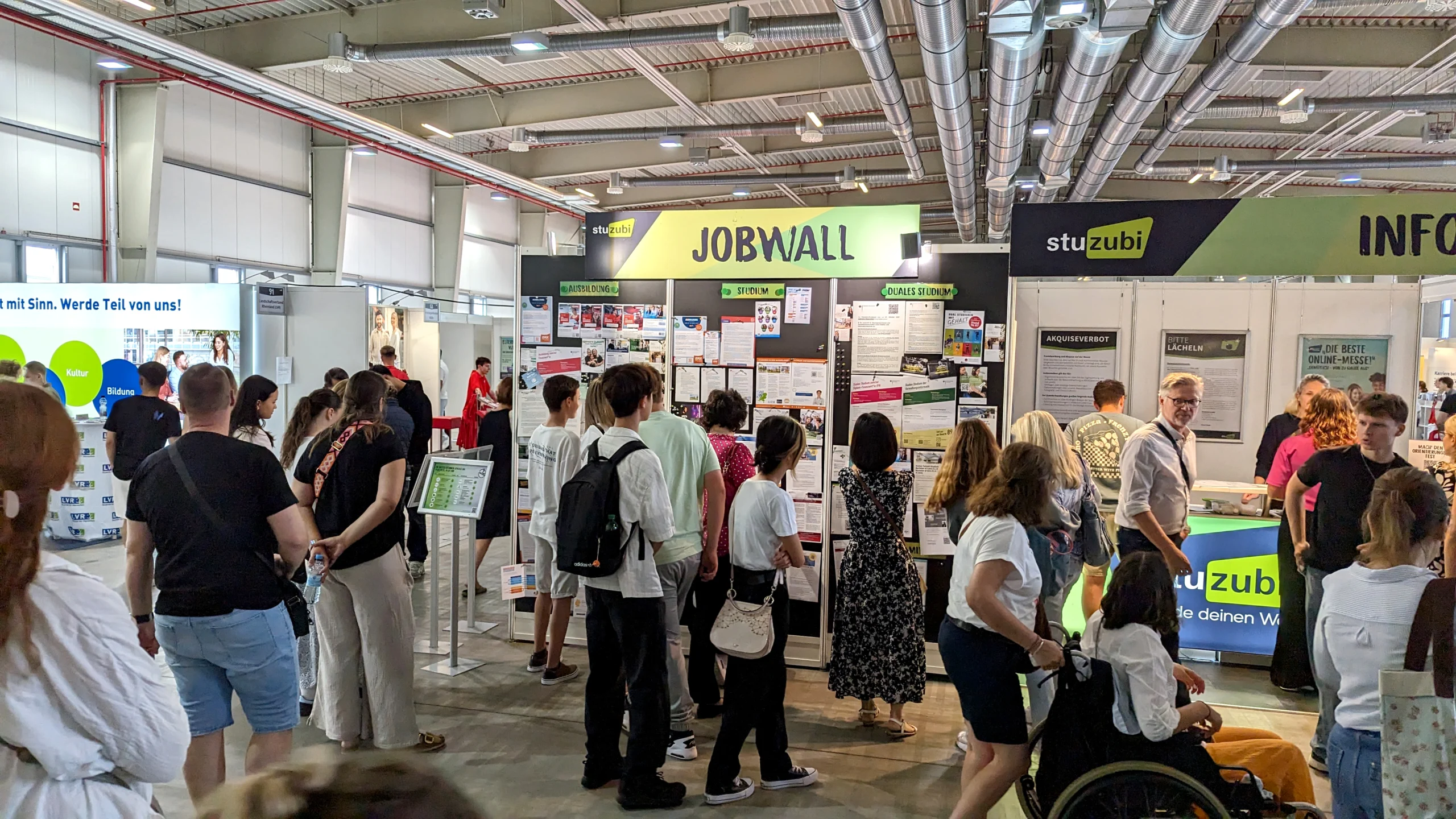 Besucher:innen stehen vor der Stuzubi-Jobwall mit den Kategorien Ausbildung, Studium und Duales Studium; im Hintergrund sind weitere Messestände und ein Stuzubi-Infotisch zu sehen. Eine Person im Rollstuhl ist im Vordergrund zu erkennen.