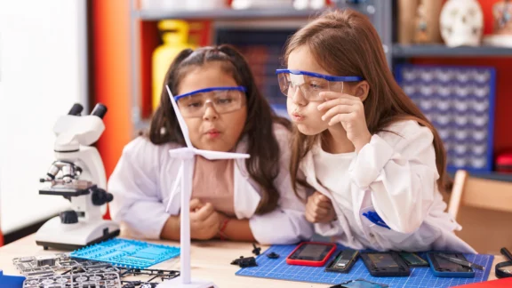 Energie zum Anfassen: Energieunterricht für die 4. Klasse Zwei Mädchen in weißen Kitteln und blauen Schutzbrillen experimentieren an einem Labortisch mit Mikroskop und wissenschaftlichen Geräten. Die Szene zeigt praktischen MINT-Unterricht in einer Schule.