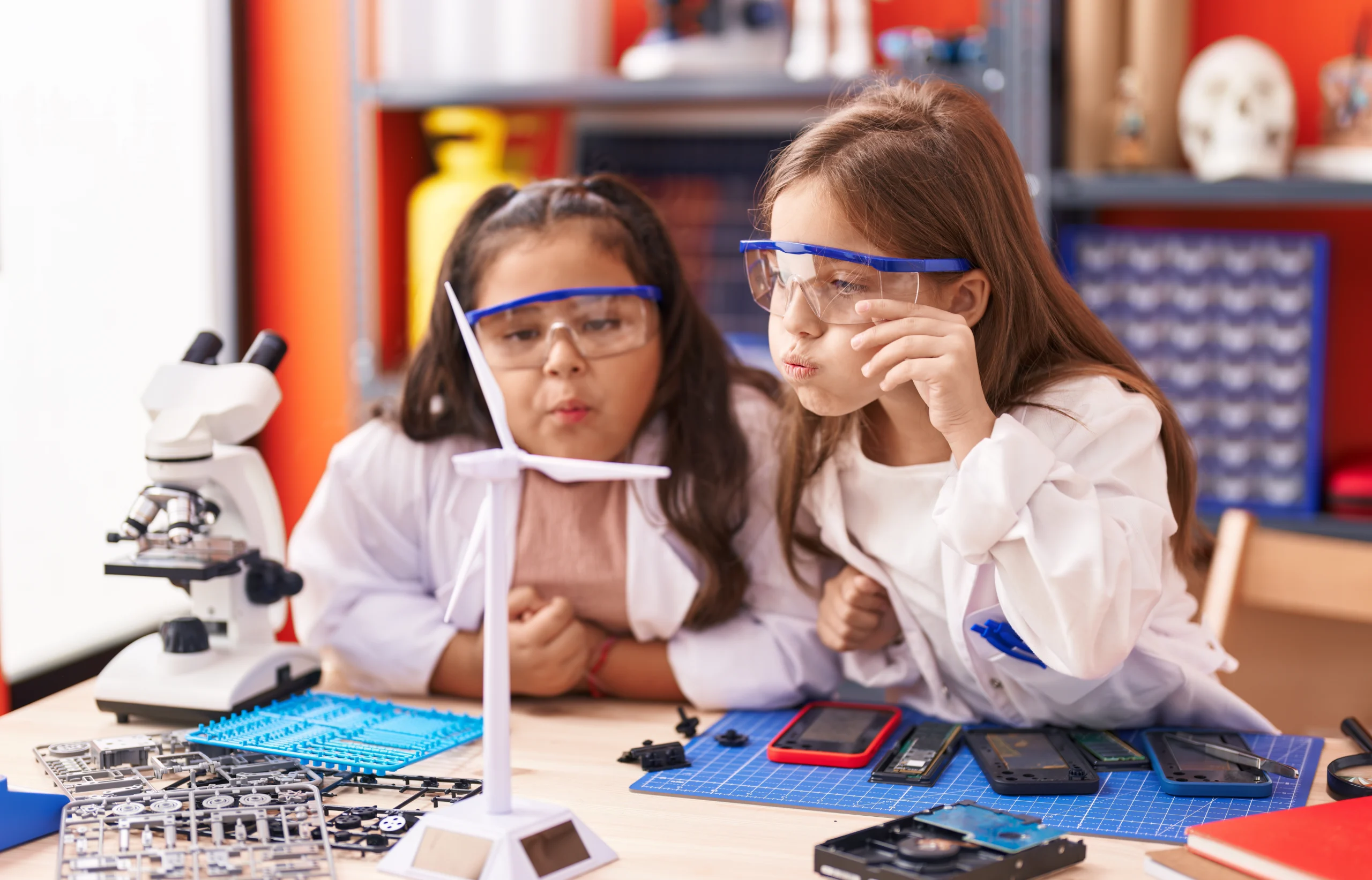 Zwei Mädchen in weißen Kitteln und blauen Schutzbrillen experimentieren an einem Labortisch mit Mikroskop und wissenschaftlichen Geräten. Die Szene zeigt praktischen MINT-Unterricht in einer Schule.