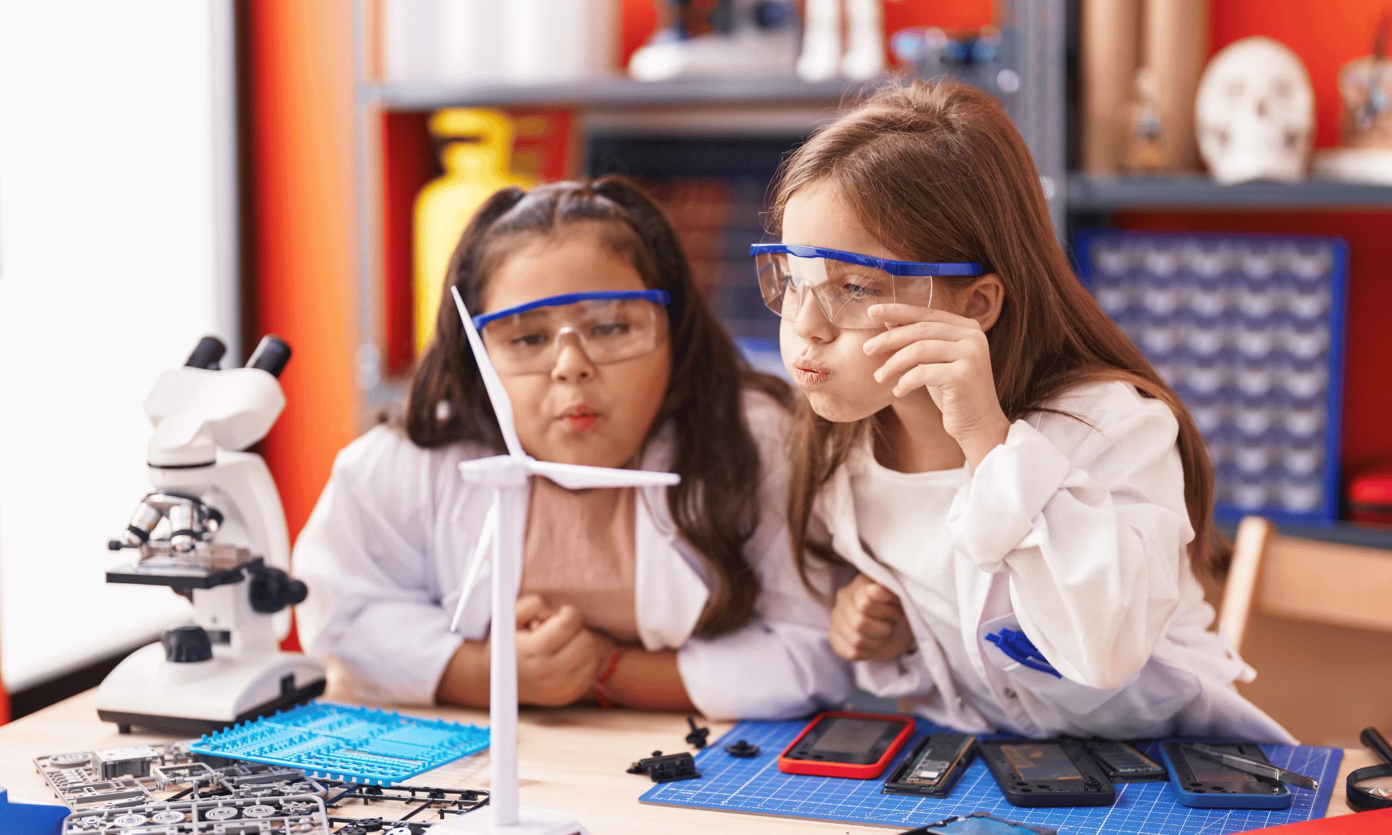 Zwei Mädchen in weißen Kitteln und blauen Schutzbrillen experimentieren an einem Labortisch mit Mikroskop und wissenschaftlichen Geräten. Die Szene zeigt praktischen MINT-Unterricht in einer Schule.