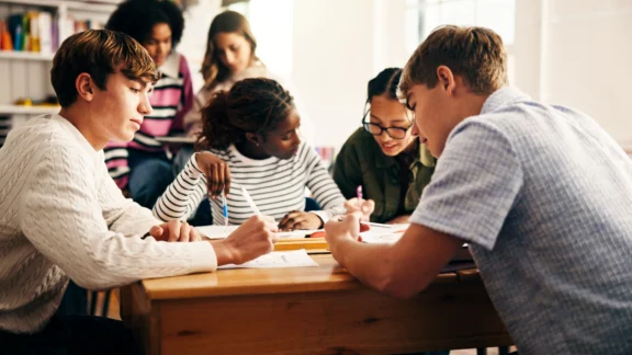 Mehrere Jugendliche unterschiedlicher Herkunft arbeiten gemeinsam an einem Holztisch in einem Klassenraum. Sie beugen sich über Hefte und Arbeitsblätter und schreiben oder diskutieren miteinander. Im Hintergrund sitzt eine weitere Schülerin und liest. Das Bild vermittelt eine konzentrierte, kooperative Lernatmosphäre.