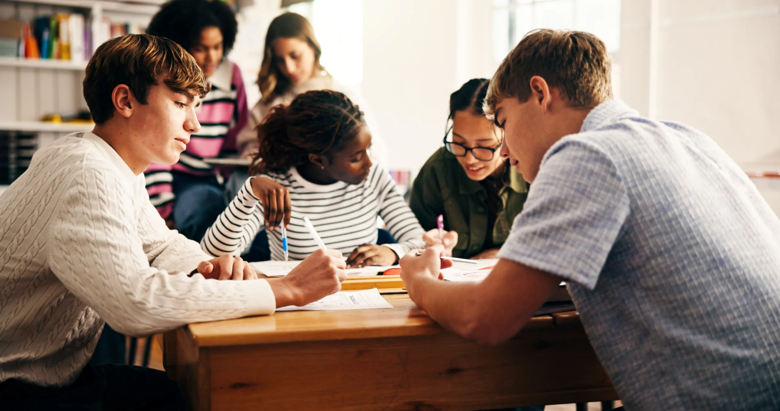 Mehrere Jugendliche unterschiedlicher Herkunft arbeiten gemeinsam an einem Holztisch in einem Klassenraum. Sie beugen sich über Hefte und Arbeitsblätter und schreiben oder diskutieren miteinander. Im Hintergrund sitzt eine weitere Schülerin und liest. Das Bild vermittelt eine konzentrierte, kooperative Lernatmosphäre.